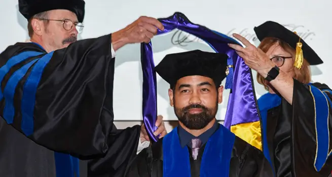 Three people in cap and gown, professors putting sash on student.