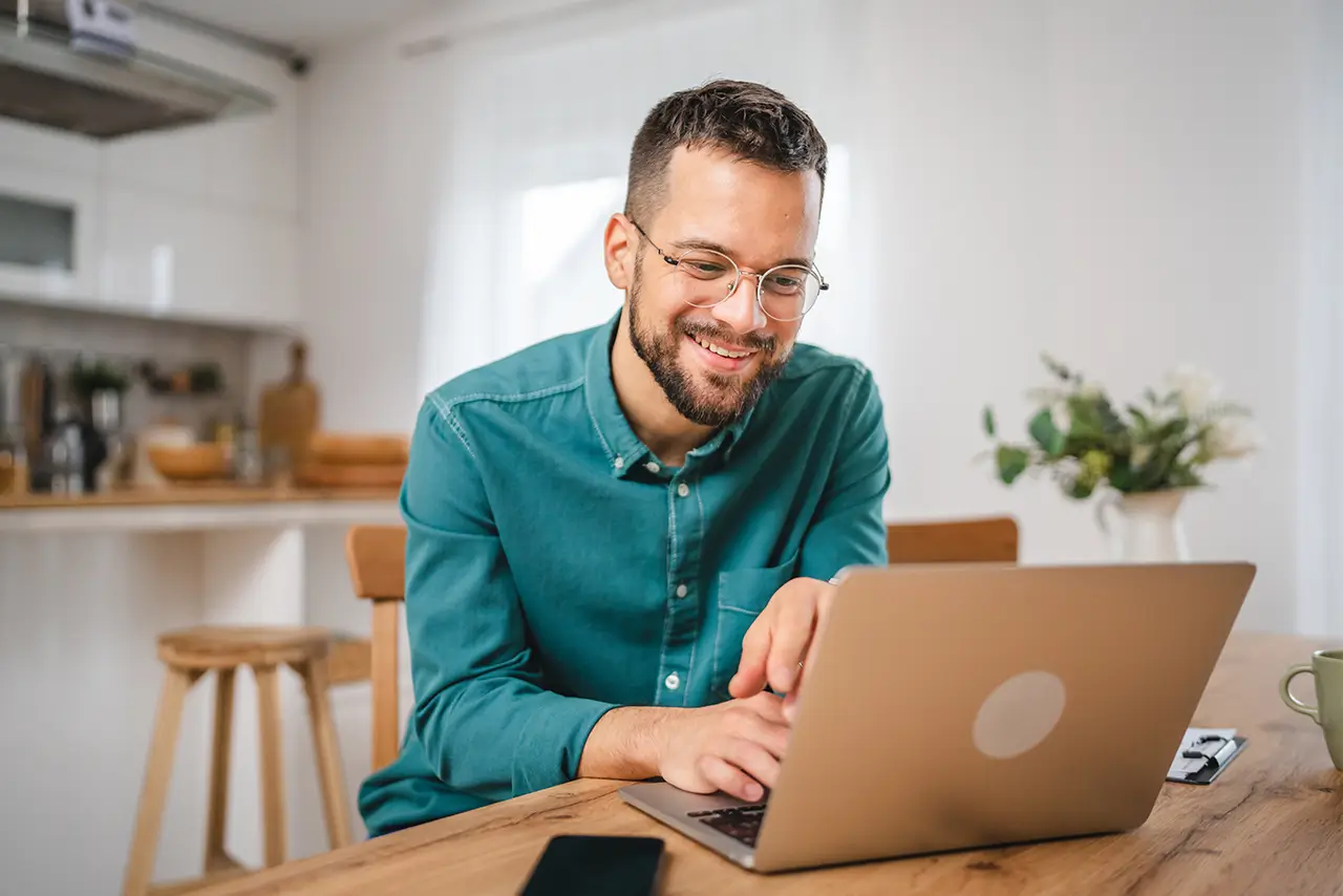 A happy man in a blue shirt sitting at a kitchen table using a laptop